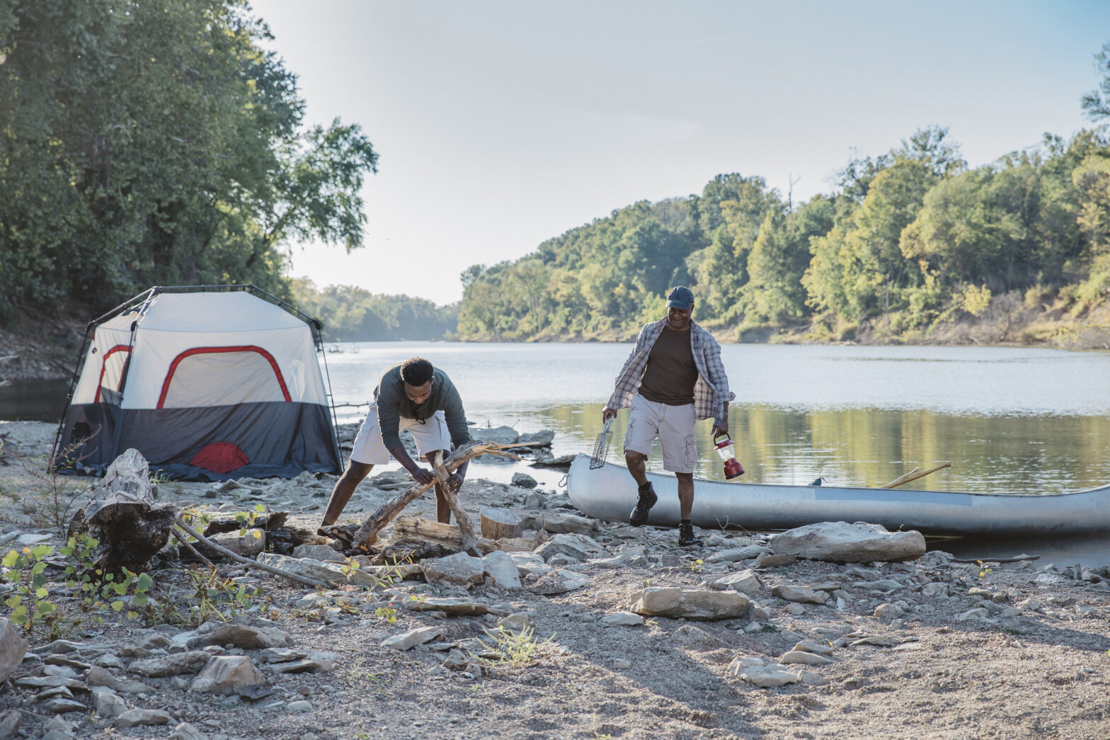 Two men set up camp beside a river—one gathers firewood while the other carries supplies from a canoe, with a tent pitched nearby and dense forest in the background.